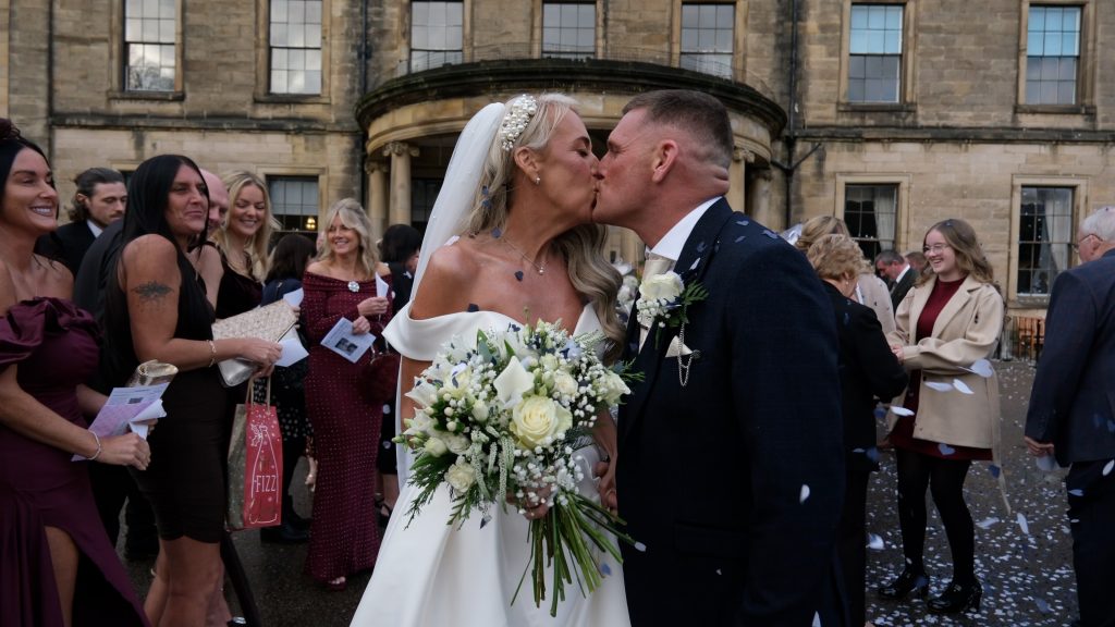 Bride and groom sharing a kiss outside Beamish Hall as guests throw confetti, capturing a joyful wedding moment at a historic North East wedding venue